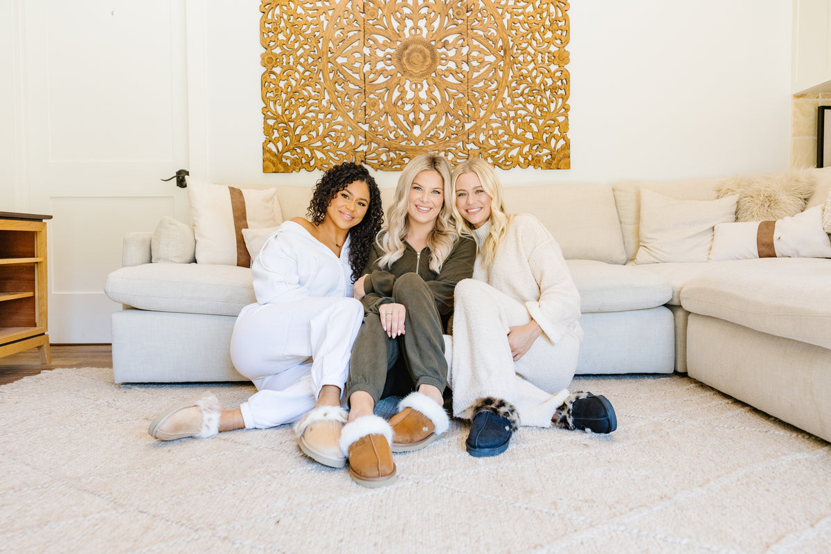 Three women sitting together on a light beige sectional sofa in a bright living room, wearing cozy lounge sets and PAWJ slippers. A large wood decorative wall piece hangs behind them, and a white rug covers the floor.