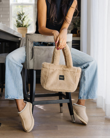 Person sitting on a chair holding a tan faux fur handbag, wearing matching shoes.